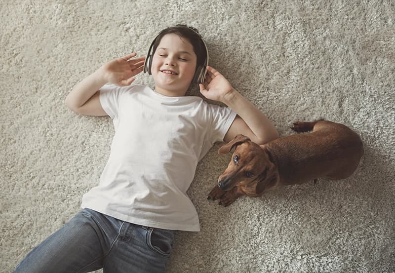 A young boy is smiling and lying on his back on the carpeted floor with his headphones on. A small dog is lying next to him and is looking at the camera.